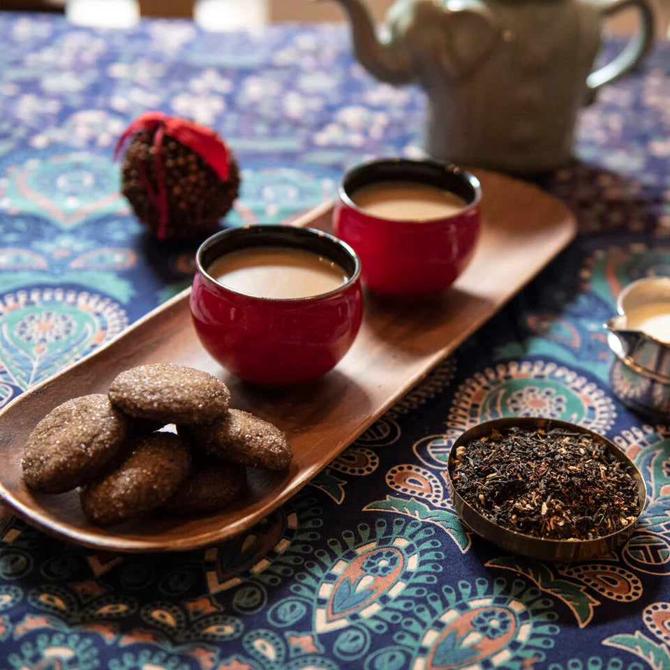 Noteworthy Selections Brewed Cacao Chai Loose Leaf Tea with milk on a wooden serving platter with two red mugs of chai next to ginger snap cookies coated in sugar on a tablecloth with a blue, green, white, and coral flower pattern. With a tin of looseleaf tea sitting on the table.