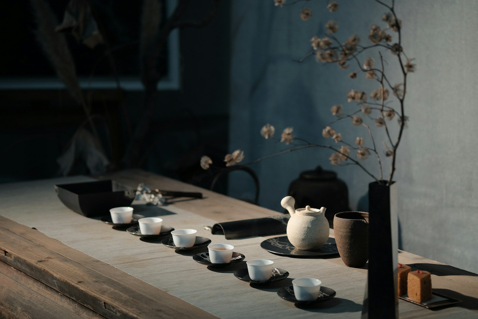 Noteworthy Selections photo of a traditional tea ceremony on a wooden table with a white porcelain tea pot, a vase of flowers, and six white serving cups with hot tea