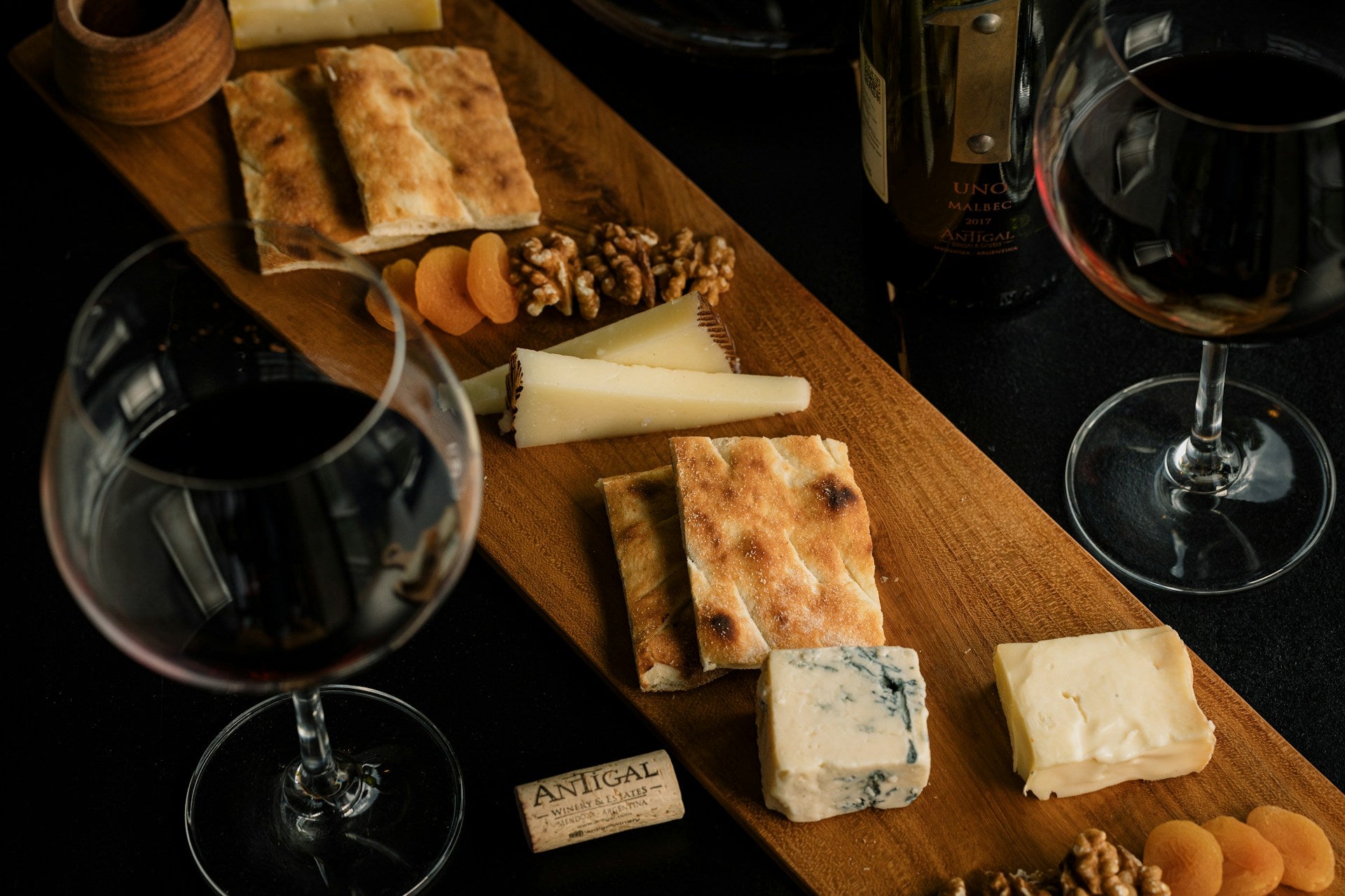 Noteworthy Selections Wine and Cheese on wooden serving board alongside two glasses of red wine, nuts, dried fruit, and slices of bread