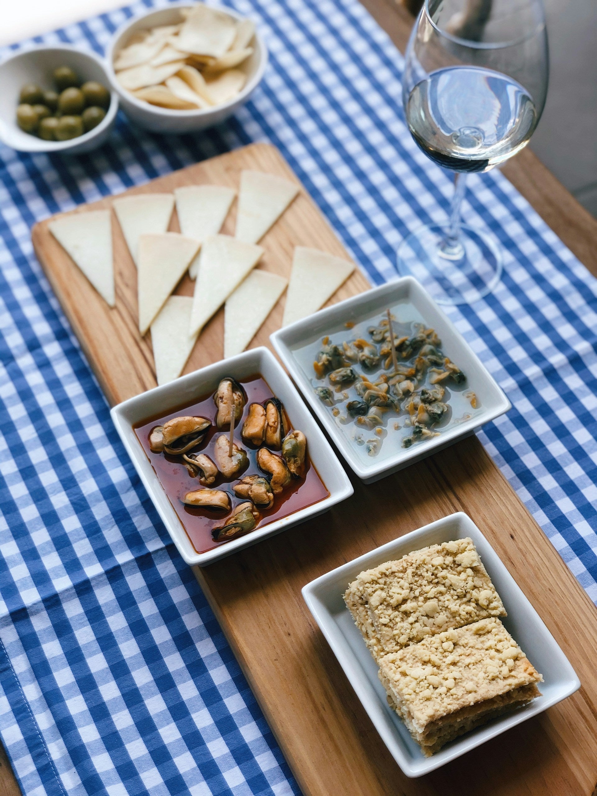 Conservas being served in small plates on a blue and white checker table cloth alongside a glass of white wine, olives, and crackers on a wooden table.