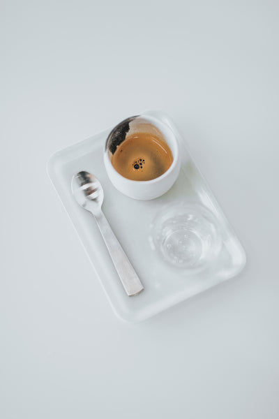 Espresso in a white cup on a white serving platter next to a glass of water and silver spoon with a white background.