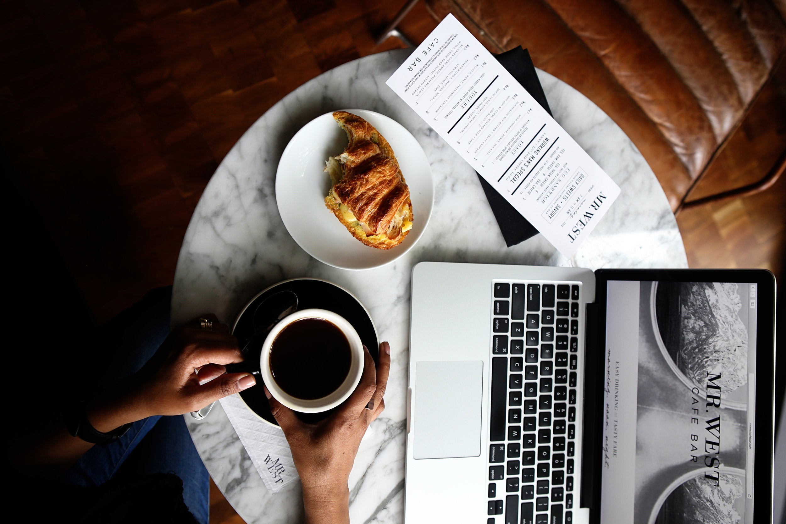 Two hands holding a mug of black coffee at a cafe table with a half eaten crossaint sandwich, receipt, and laptop open while working on staff picks for the website.