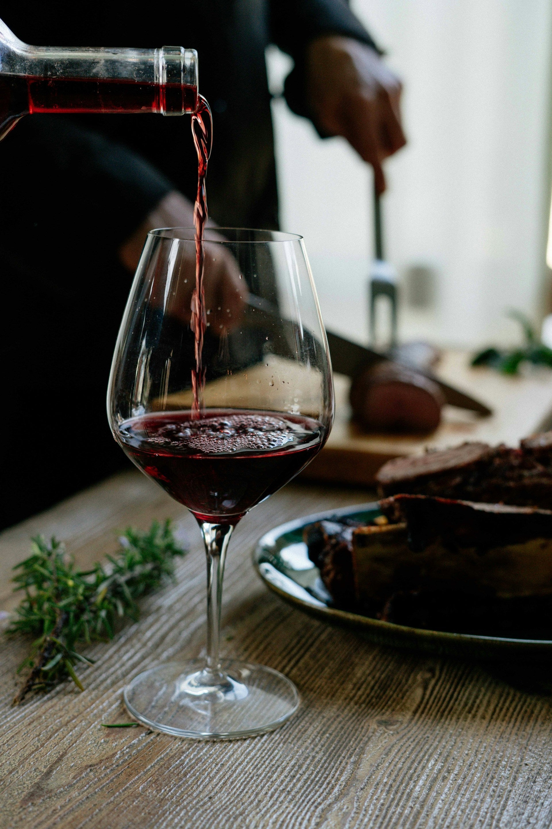 Wine being poured into a glass sitting on a wooden table while meat is being carved in the background.