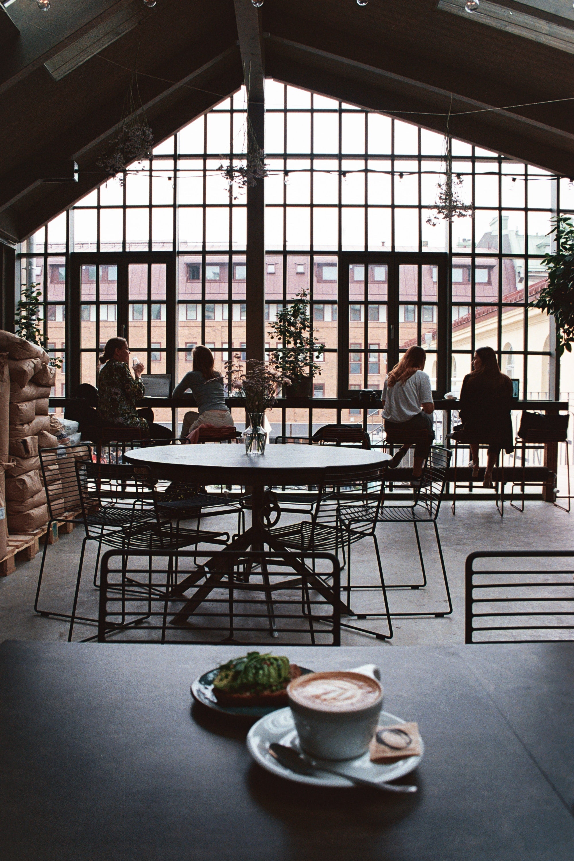 A latte in a white mug served with food awaits on counter for pick up at american roasters cafe. While people sit in the background looking out the windows, talking and sipping coffee.