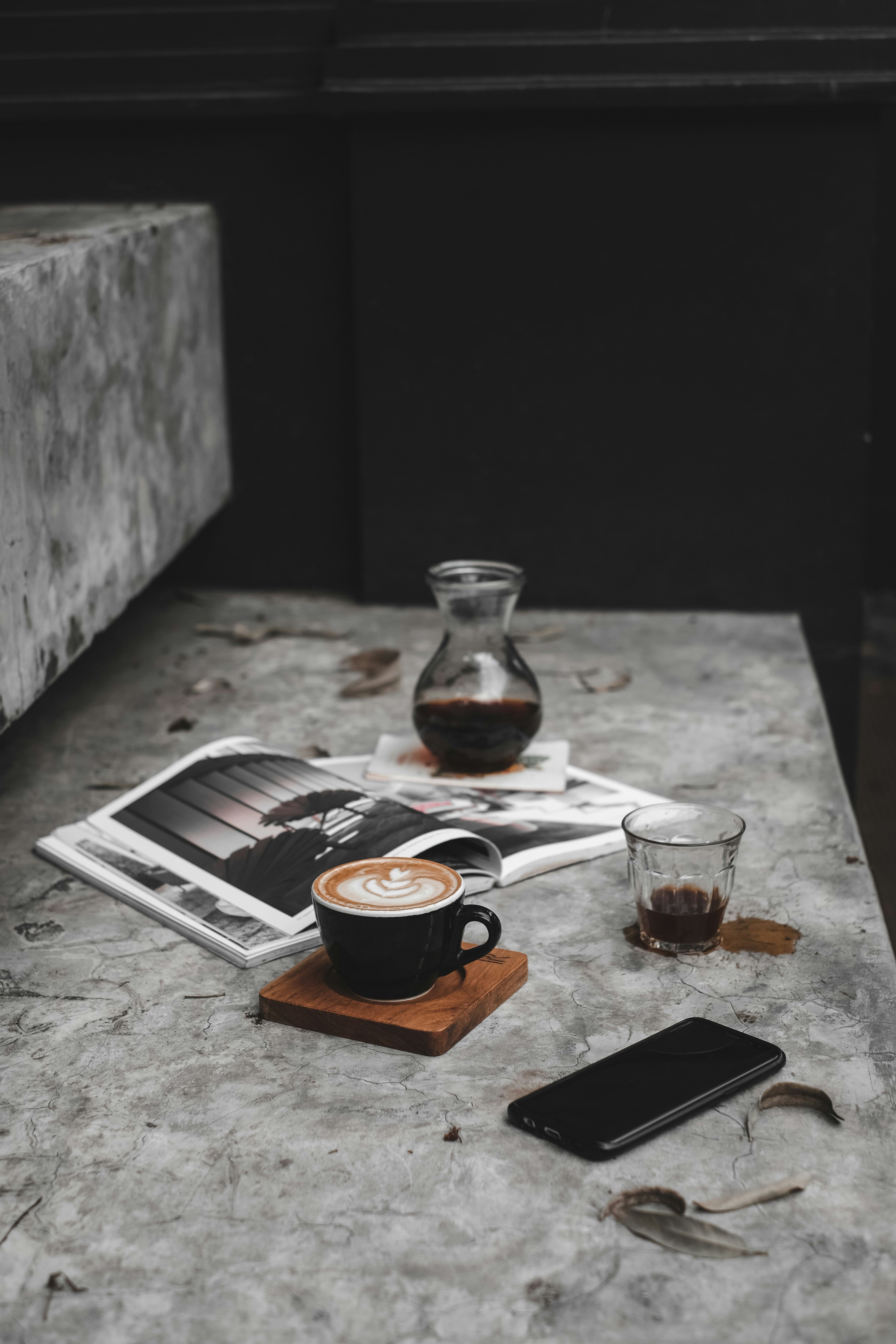 A mini carafe of black coffee placed on a magazine, a glass of black coffe with spilled coffee around it, a mobile phone, and a latte in a black mug with latte art on top sitting on a wooden square all along a gray marble staircase.