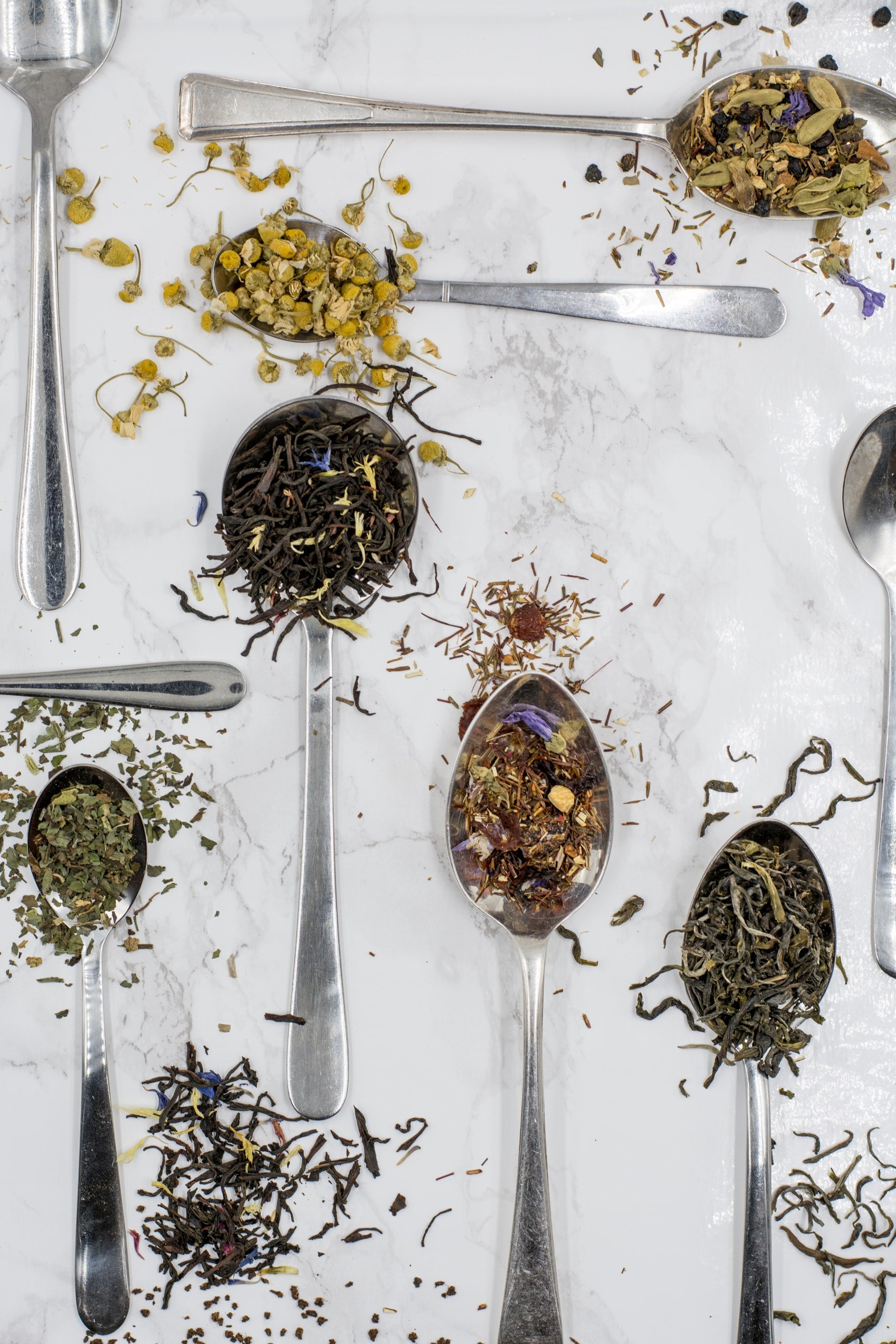 A variety of single origin tea in spoons lying on a white table cloth with loose leaf tea leaves sprinkled about.