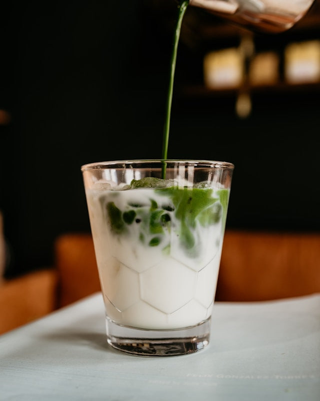 Noteworthy Selections Matcha being poured into a glass full of milk for a matcha latte. Sitting on a white table.